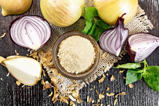 Onion powder in a bowl on burlap, purple and yellow onions, dried onion flakes and basil on wooden board background from above